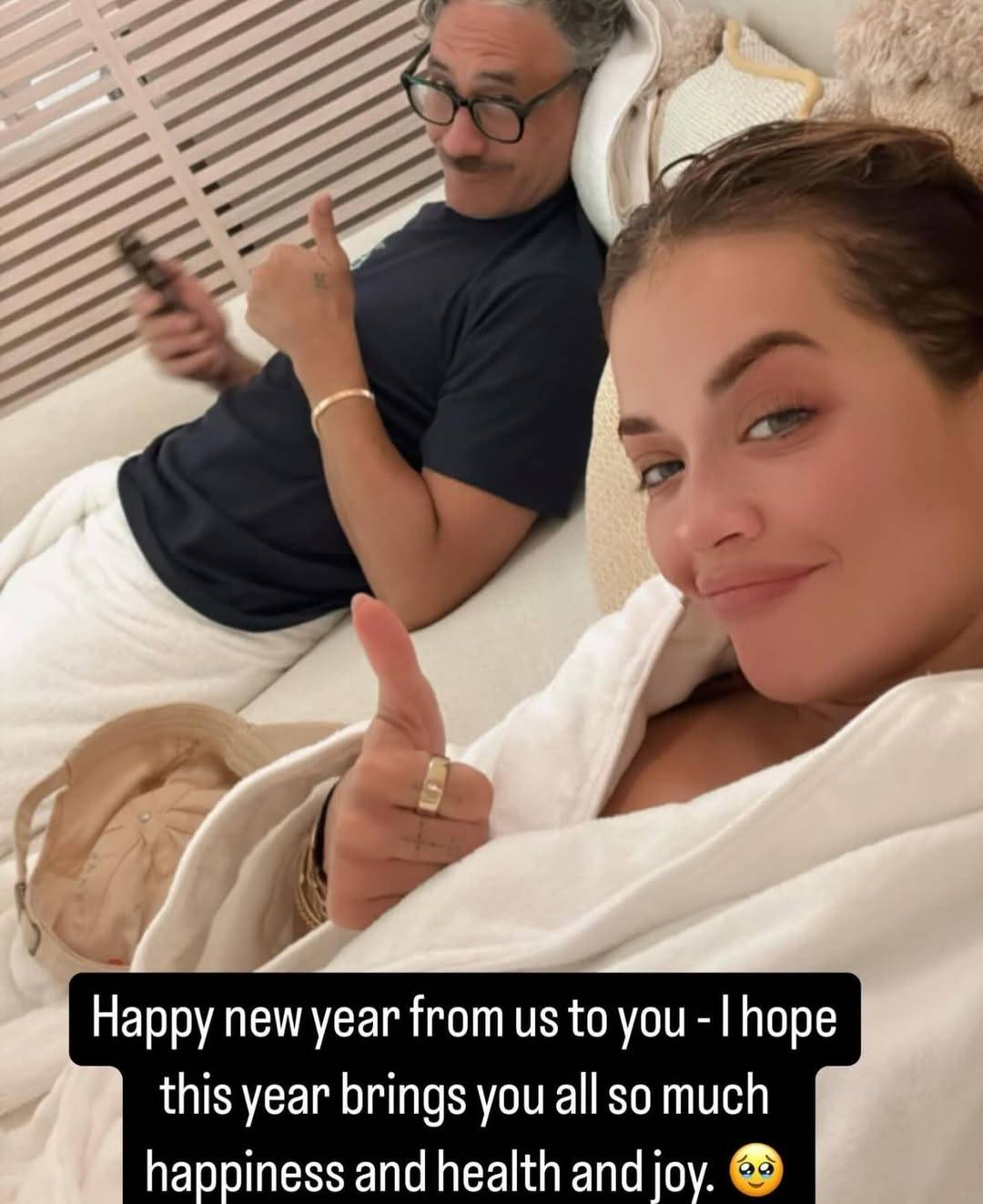 Selfie taken by Rita Ora with her in the foreground and Taika in the background. She and Taika are lying back on a futon, smiling and giving thumbs up. Taika is holding his phone and looking at the camera over the rim of his glasses. Rita is wearing a fluffy white bathrobe, while Taika is in a dark blue t-shirt and a white towel. Text at the bottom reads: ‘Happy new year from us to you – I hope this year brings you all so much happiness and health and joy (holding back happy tears emoji)’.