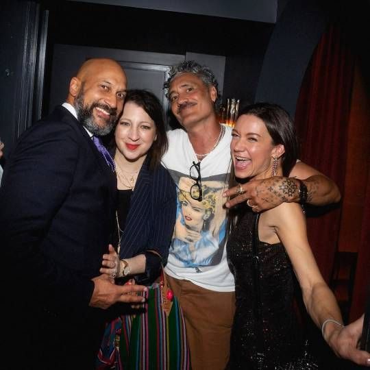 Photo of Taika and three friends at a party. They stand with their arms around each other, smiling at the camera except for one of the women, who is laughing and looking at something off to the side.