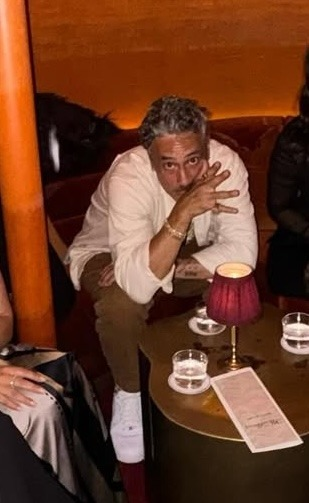 Taika sitting with others at a low table. The photo is cropped so only he is fully visible. He is leaning forwards with his elbows on his knees and looking up at the camera, one hand held up in front of him, his fingers spread wide to hide most of the bottom half of his face. On the table sit a small lamp, a menu and three glasses containing clear liquid.