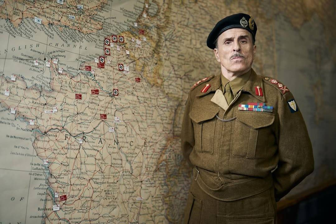 Con wearing an olive green military uniform with many different badges and a black beret, looking sternly at the camera with his hands behind his back. He is standing in front of a WW2 era map of Europe, with France, Belgium, the English Channel and the southern coast of England visible. Small pieces of paper have been placed on the map, with a number of swastikas in the north-east of France marking areas occupied by the Nazis.