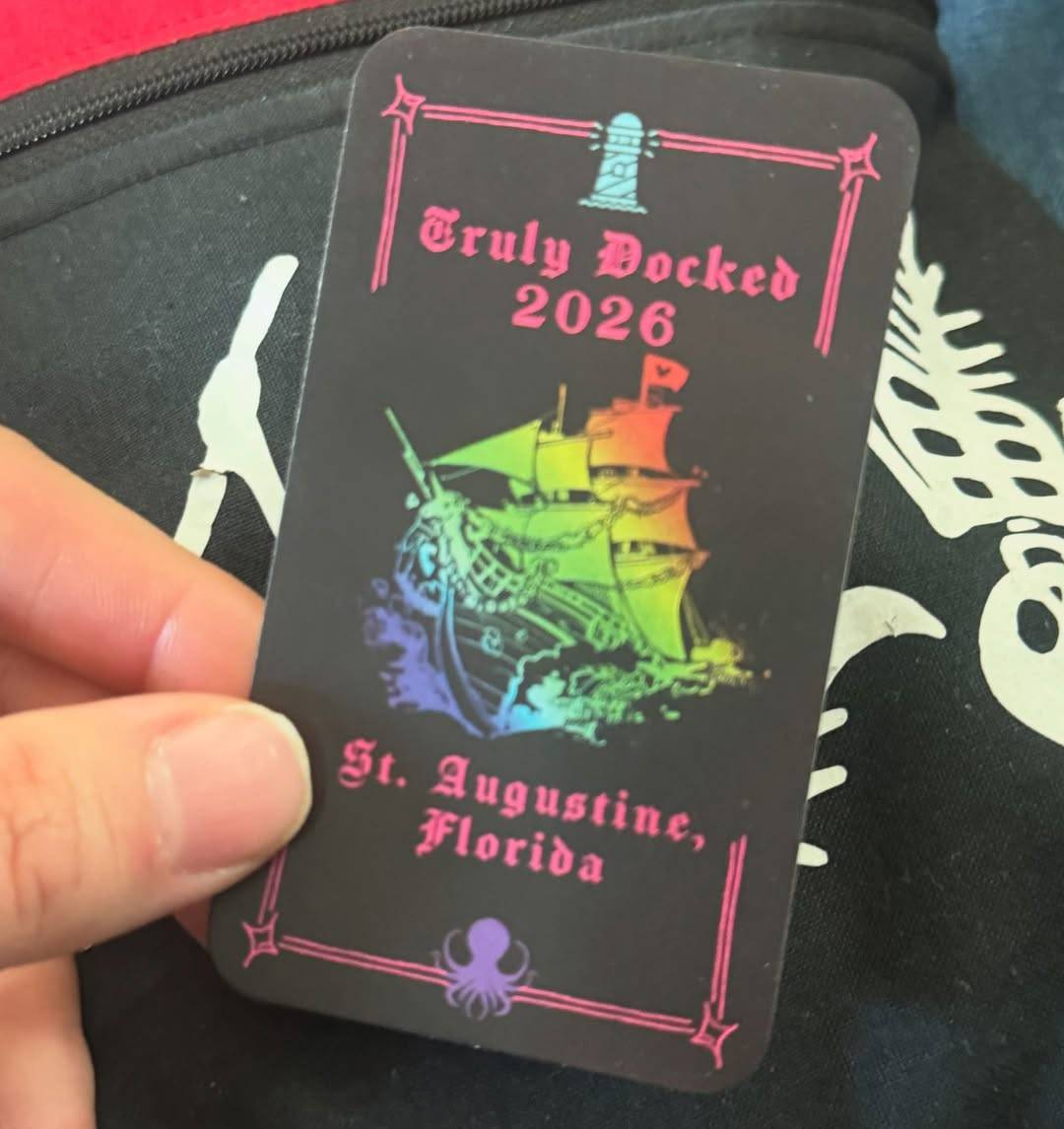 Photo of a small black card with an image of a sailing ship in rainbow colours parting the waves at the centre, 'Truly Docked 2026. St. Augustine, Florida' written in hot pink Gothic type above and below. At the top and bottom of the card, there is a drawing of a small teal lighthouse and a purple kraken respectively, and thin hot pink lines with a little star shape in each corner form a partial border near the edge of the card.