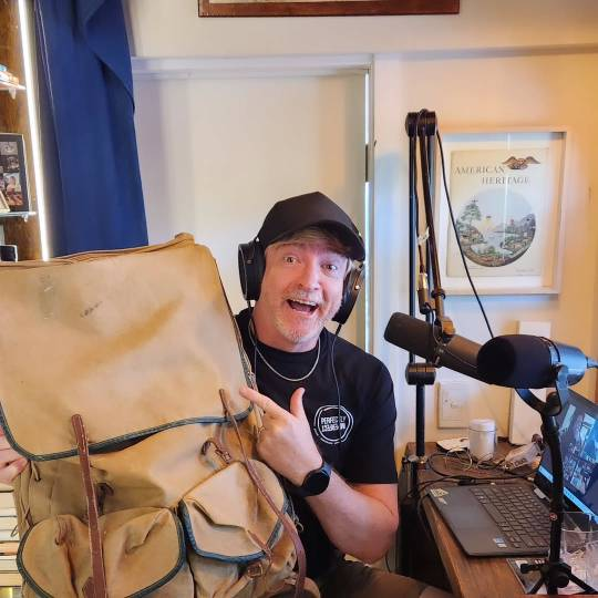 Photo of Rhys smiling excitedly, his mouth open and eyes wide as he holds up a large old canvas backpack, pointing at it with one hand. He is sitting at a desk with a laptop and two large microphones, and is wearing a pair of over-ear headphones.