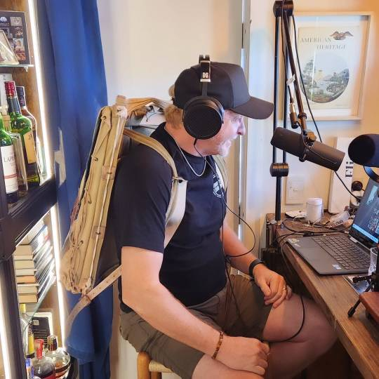 Another photo of Rhys sitting at his desk while recording the podcast. He is now wearing the backpack, which looks flat and blocky when empty, and he is grinning down at his laptop screen.