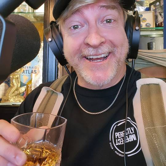 Selfie of Rhys with his backpack and headphones on, looking delighted, with a big, open-mouthed smile on his face. In celebration of receiving the backpack, he is holding up a short, square-shaped glass filled to a third with amber liquid.