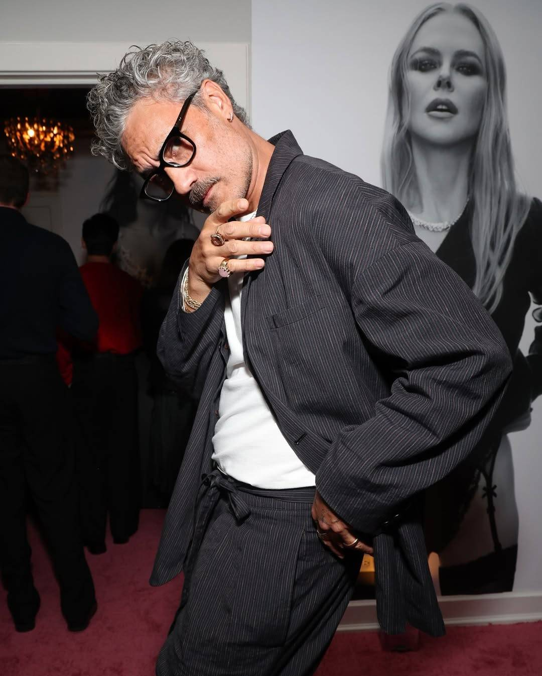 Photo of Taika, seen from the side from about the thighs up. He is wearing a baggy dark pinstripe suit, a white shirt, glasses with a thick black rim, and several chunky rings on his fingers. He is looking sidelong at the camera and striking a pose with his head bent slightly, one hand raised to near his chin and the other hand pulling back part of his jacket to better show off his trousers, which have a matching fabric belt that is tied into a bow at the front. 