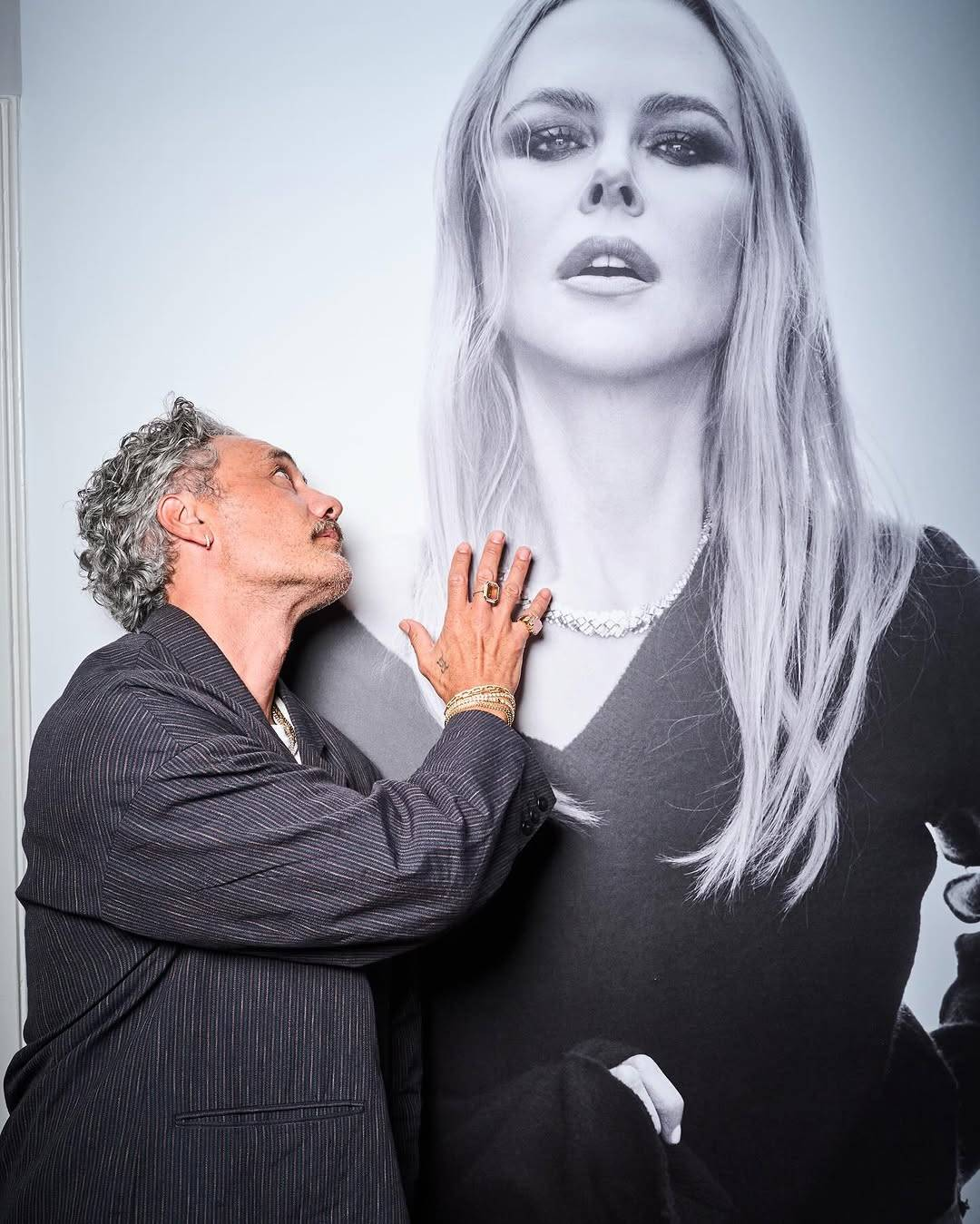 Taika, dressed in the suit from the previous picture, stands in front of an oversized black & white photo of Nicole Kidman that fills the entire frame of this image. He is leaning on the wall with one shoulder and looking up reverently at Nicole's face, with one hand gently placed on the spot where her long blond hair falls over her shoulders.