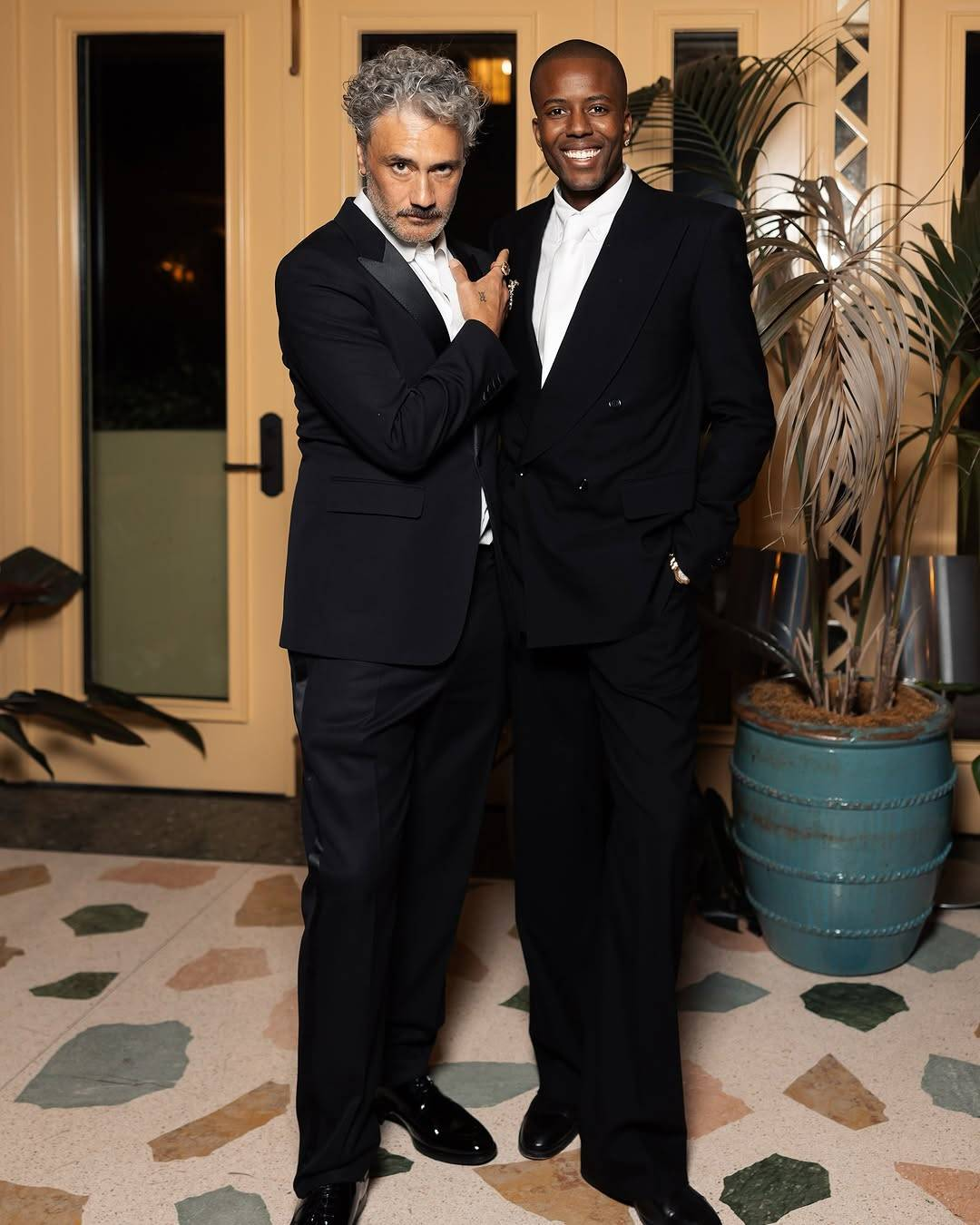 Full-body photo of Taika and Vas J Morgan, both dressed up in dark suits, white dress shirts and polished black shoes. Vas's suit is black and he is wearing a white tie, while Taika's is a very dark blue, worn without a tie. Vas is smiling broadly at the camera with his hands in his pockets, while Taika is looking up at the camera with his head slightly bowed and a serious expression on his face, one hand pointing appreciatively at Vas.