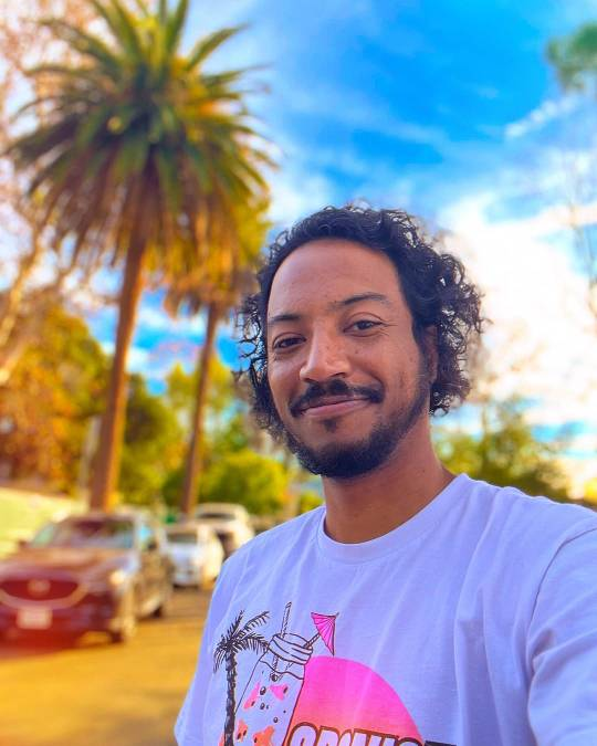 Selfie of Samba, wearing a white Spanish Jackie's shirt and smiling at the camera. Behind him, there is a street lined with palm trees, and the sky above is a vivid blue streaked with wispy white clouds.