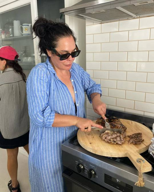 achel House stands in a kitchen, cutting slices off a large piece of cooked meat on a wooden cutting board, which is sitting on top of a stove. She is wearing sunglasses and a light blue shirt dress with white stripes, her hair up in a messy bun.