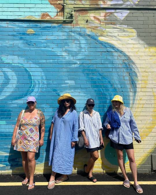 Rachel House and three of her friends lean against a colourfully painted exterior brick wall. She is wearing the striped dress and sunglasses from the previous image, now with her hair down and paired with a yellow sun hat and a pair of sandals. One of her friends is wearing a colourful mini dress with knotted shoulder straps, and the other two are both wearing baggy blue & white button-up shirts and dark shorts. They are each wearing a hat and sunglasses in different styles