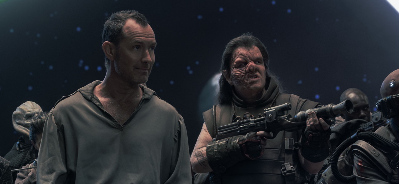 Dominic as Beef in Star Wars Skeleton Crew, standing next to Jude Law as Jod Na Nawood, seen from about the waist up. Beef is pointing a rifle at someone off camera. One side of his face his heavily scarred, his nose squished and his top lip on that side pulled up to reveal his teeth. He has long dark hair, which is brushed back and falling down over his shoulders. He is wearing a vest that looks to be made of leather, with a stiff collar protecting his neck. His hands and forearms are clad in heavy black gauntlets. Jod Na Nawood is smiling placidly next to him, is eyebrows raised and brow furrowed. Next to and behind them, other characters, also wearing armor, are partially visible.