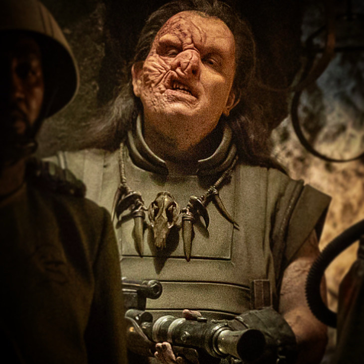 Close-up of Dominic as Beef, again seen from the waist up. In addition to the armor from the first image, he is also wearing a black beaded necklace with an animal skull, large canine teeth and claws on it. He is holding the same rifle, but only part of it is visible in this image.