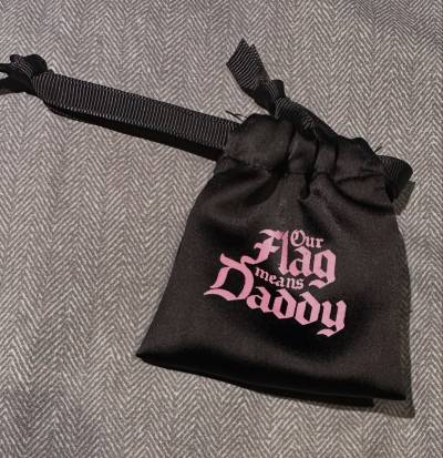 A small black pouch with 'Our Flag means Daddy' written on it in pink, in the same font as the OFMD logo. 