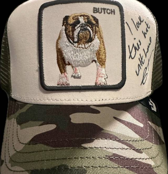 A baseball cap with an English bulldog and the word 'butch' embroidered on it. The main part of the hat is a light beige and the visor has a camouflage pattern. Handwritten in black marker on the side of the cap are the words 'I love this hat, with love (Con's signature).' 