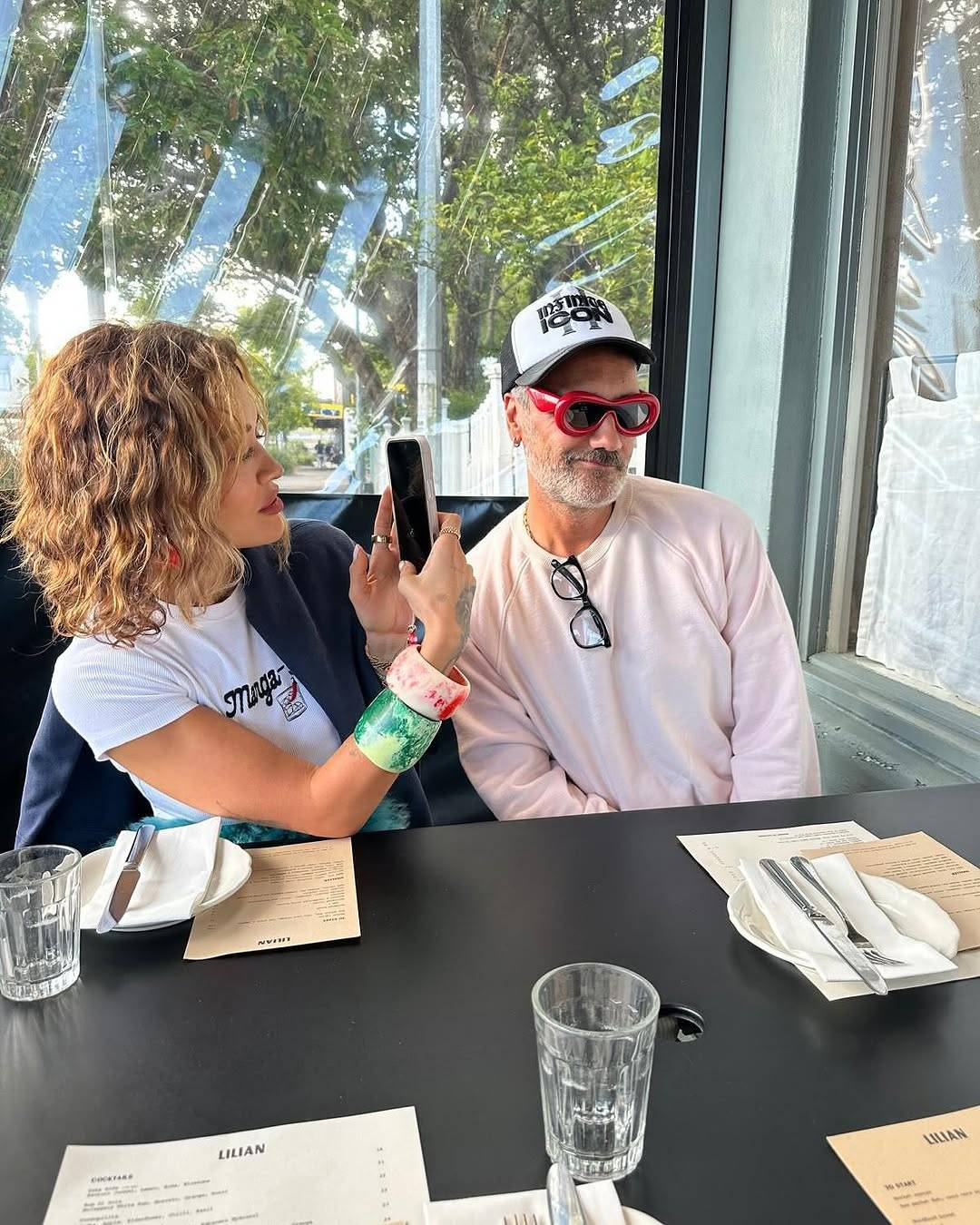 Rita Ora and Taika Waititi sit at a restaurant table. Taika is wearing a pair of thick-rimmed red sunglasses that almost resemble ski goggles in shape. His usual black pair of glasses is tucked into his collar. Rita, who is sitting next to Taika, is taking a photo of him on her phone, and he is leaning towards her with a lopsided grin, his face tilted slightly away from her.