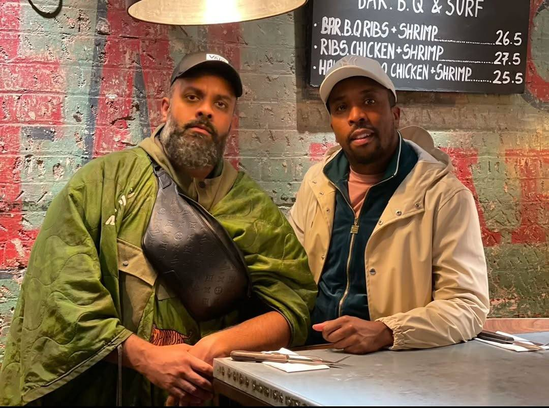 Guz and Kiell stand at a tall table or counter in a fast food restaurant. They are both leaning on the table with one elbow. Guz is looking at the camera with a serious expression, while Kiell is smiling slightly.