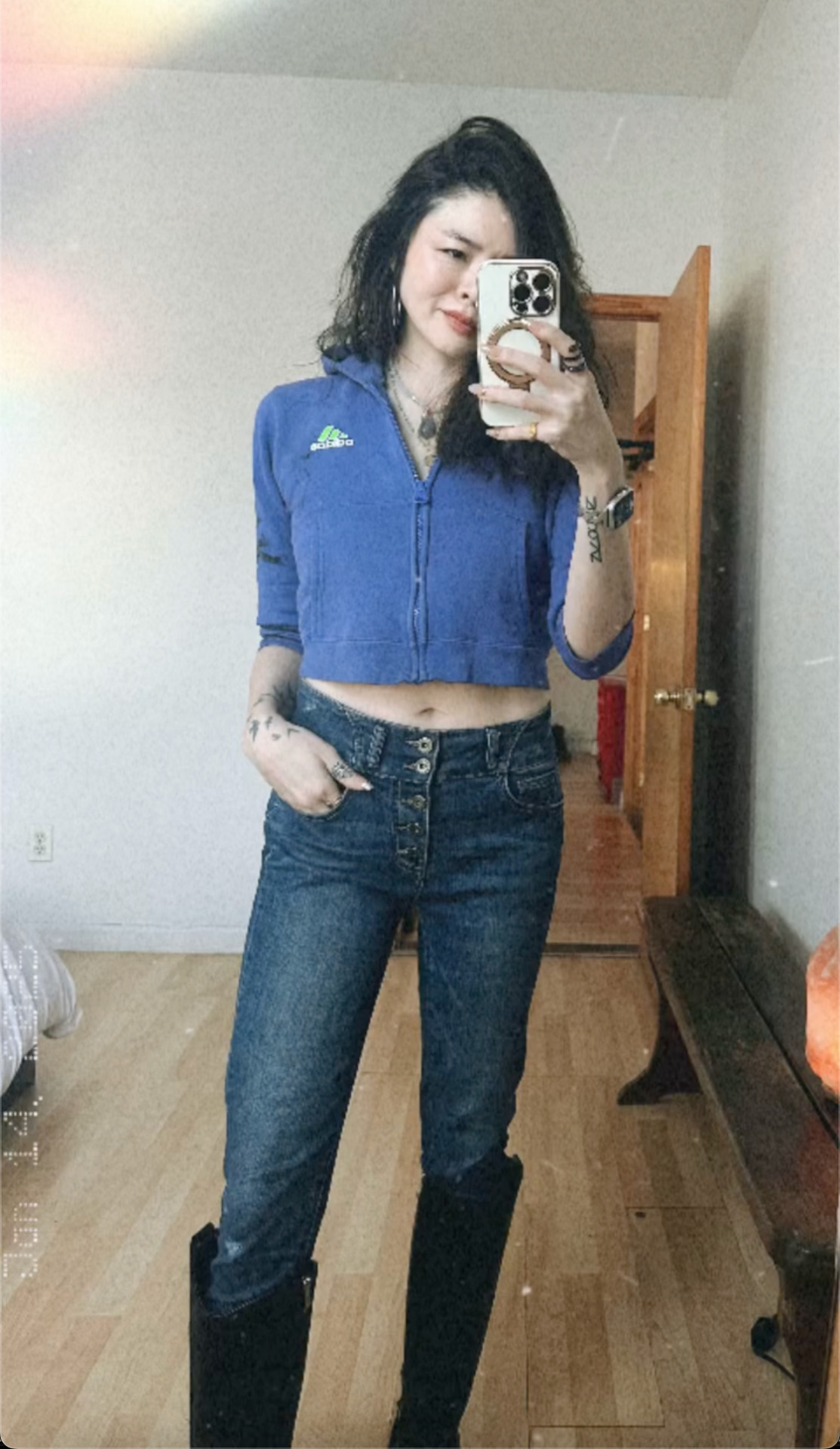 Mirror selfie of Ruibo wearing a cropped blue hoodie, blue jeans and knee-high black boots. A sliver of belly is showing between her jeans and her hoodie. She is smiling slightly and looking at her phone, which she is holding up to take the picture. The fingers of her other hand are tucked into her jeans pocket.