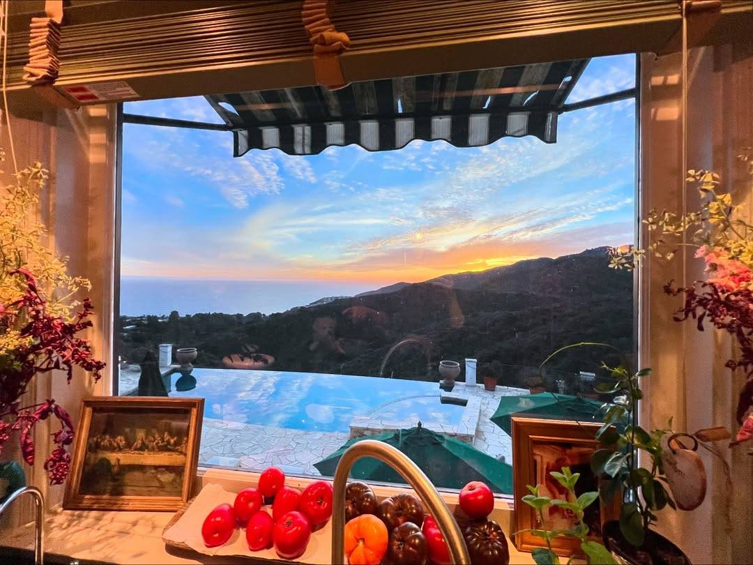 Photo of a sunset shot through a kitchen window. Just outside the window, there is a patio or tiled backyard with an infinity pool looking onto wooded hills and the ocean in the distance. The sky is a bright blue streaked with fluffy white clouds, the horizon painted orange and yellow by the setting sun. On the inside window sill sits a plate of red, orange and purplish tomatoes, flanked by two small framed paintings. Two bouquets of flowers are partially visible standing on either side of the window.