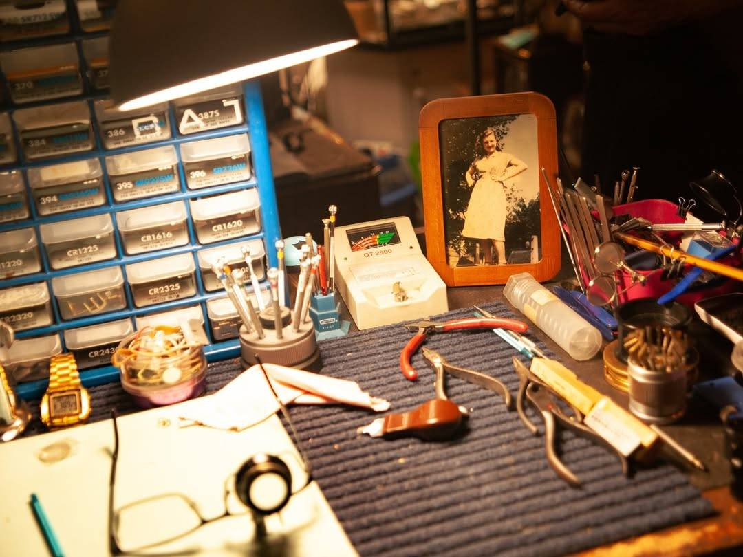 A watchmaker's workbench decorated by Lindsey for the movie Watching Walter. The small space is filled with various watchmaking tools stored in small containers or strewn across the bench. Watch parts are sorted into clear labeled bins inside a small storage cabinet at the back. A framed photo of a woman standing in front of a tree with her hands on her hips is placed prominently in one corner of the workbench.