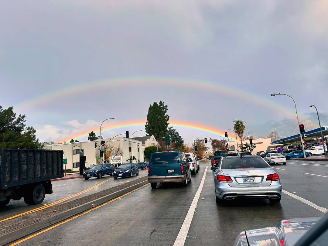 Photo of a double rainbow, shot from the window of a car on a busy road. The larger and fainter of the 2 rainbows stretches across the entire width of the image, making it seem like the road leads directly to it.