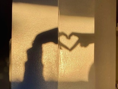 The shadow of two hands forming a heart with their pointer and middle fingers, cast onto a wall by warm yellow light.