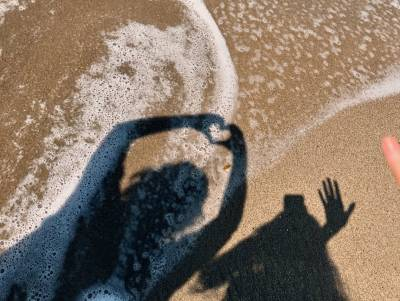 Two people's shadows on a beach, just at the edge of the water so the foam left behind by the waves is still visible. One person is holding their hands up in the shape of a heart while the other is taking the photo with one hand and waving at the ground with the other.