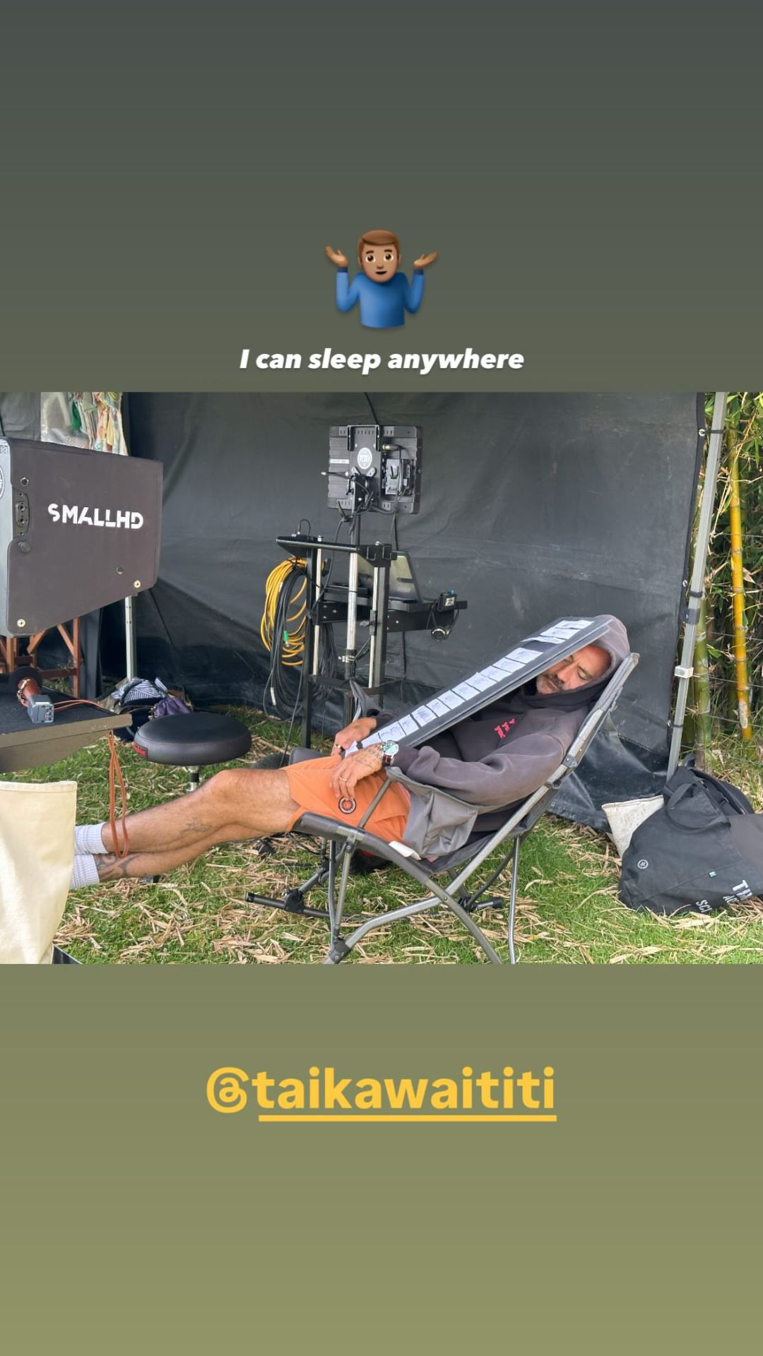 Instagram story shared by Taika: a photo of Taika sleeping in a folding rocking chair surrounded by filming equipment. He is wearing shorts and a hoodie with the hood up, his head is turned to one side, and a stiff sheet of plastic(?) covers the upper half of his body to provide some cover and block out the light. His legs are stretched out, his feet propped up on something out of frame. Below the photo, the person who originally posted it has tagged @ TaikaWaititi. Above, Taika has added a shrug emoji and the words ‘I can sleep anywhere’.