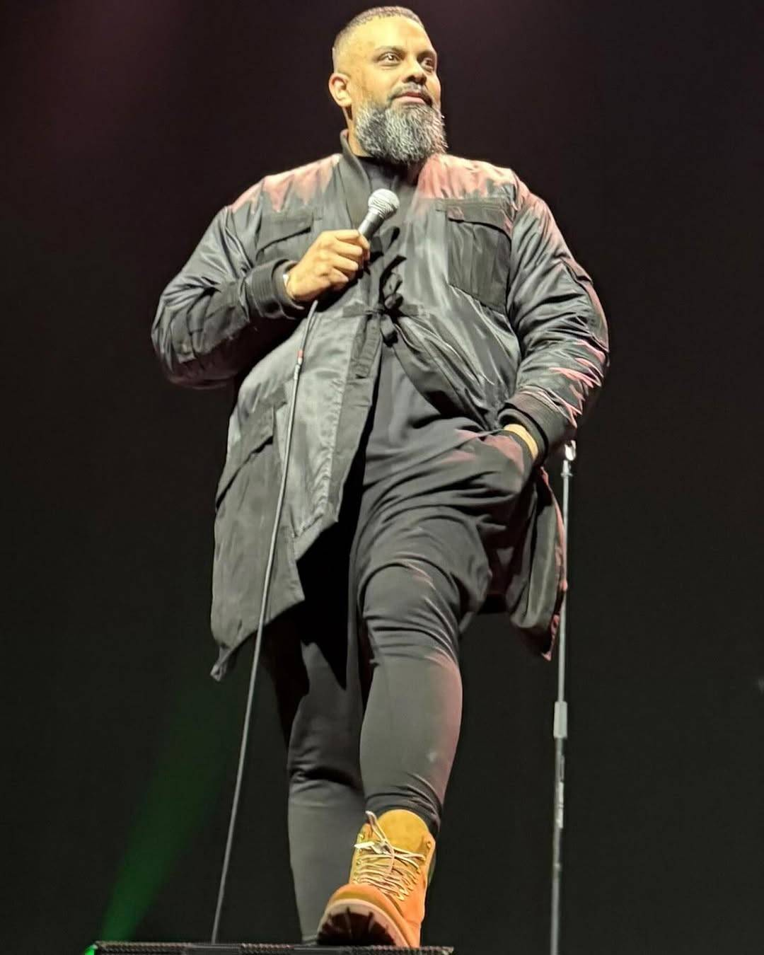 Photo of Guz on stage, shot from a low angle. He is standing with one foot up on a monitor, smiling at the audience and holding the microphone in his right hand, left hand tucked into his trouser pocket.