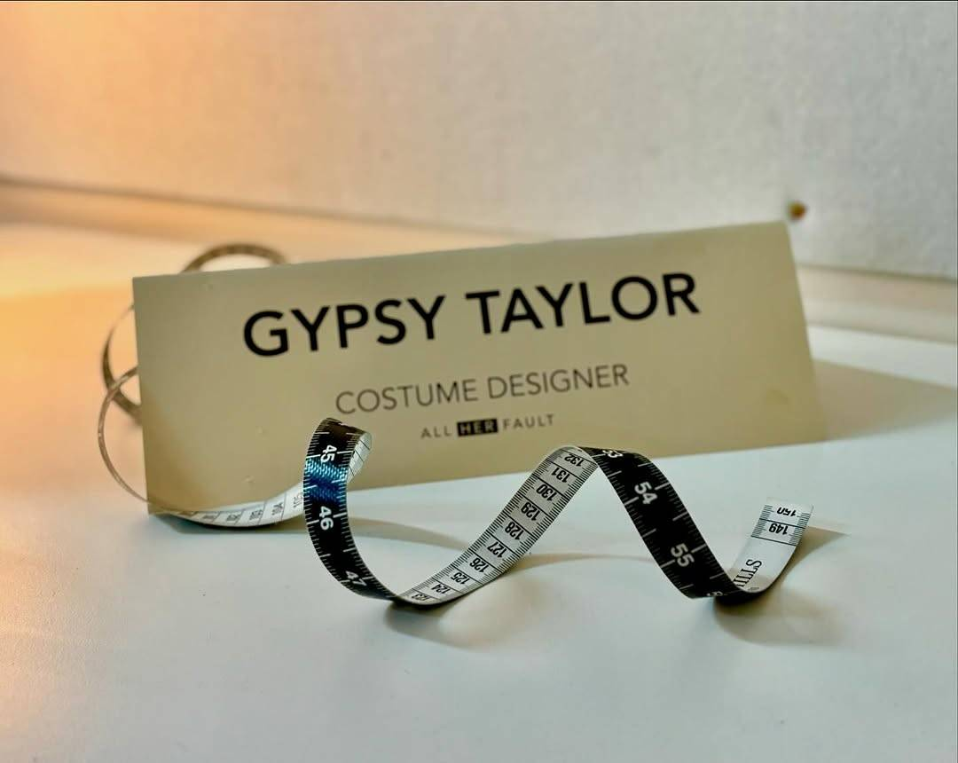 Photo of a paper name card and an unfurled length of black & white tape measure. The card reads: ‘Gyspy Taylor – costume designer, All Her Fault’.