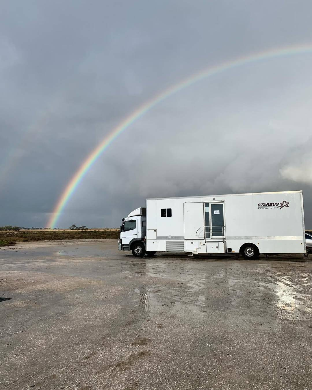 A white costume trailer parked in an otherwise empty lot. Directly above, a bright rainbow can be seen in the cloudy grey sky.