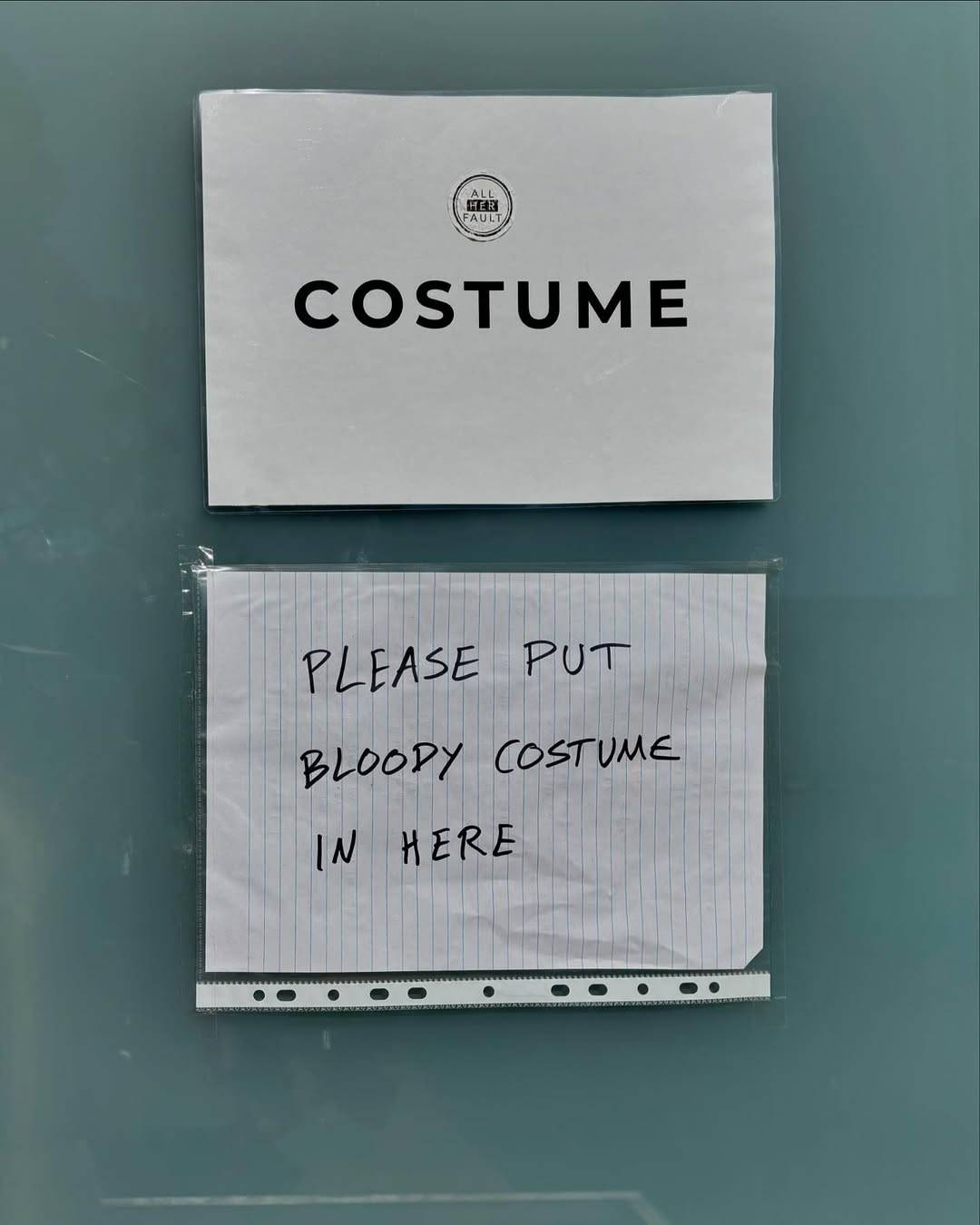 Two pieces of paper hung on a wall. One is laminated and has the ‘All Her Fault’ show logo and the word ‘Costume’ printed on it. The other one below it is handwritten and placed inside a plastic sleeve. It reads: ‘Please put bloody costume in here.’