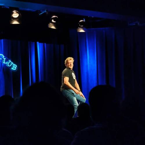 A photo taken from within the crowd at Rhys's stand-up show. Rhys is sitting on a stool in front of a blue curtain and looking at the audience as he speaks.