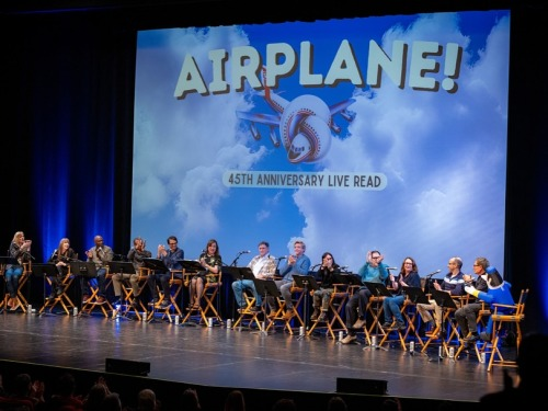 13 people sit side by side in folding chairs on a stage, with a 14th chair occupied by Otto the inflatable dummy pilot from the film 'Airplane!' Each person has a microphone and a metal lectern standing in front of them, and behind them, a large screen displays a picture of an airplane tied into a knot and the words 'Airplane! 45th anniversary live read', on a background of a blue sky with white clouds.