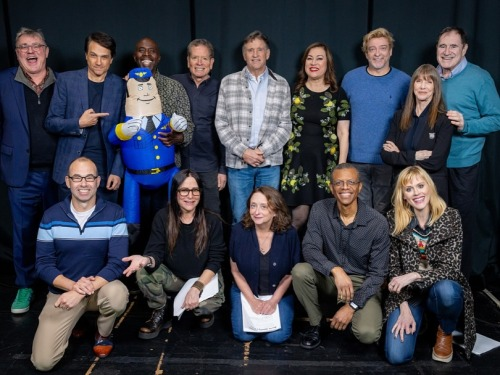 Group photo of the actors who participated in the 45th anniversary live read of 'Airplane!' 9 people, including Rhys Darby, stand at the back, one of them holding up Otto the inflatable pilot, while 5 people kneel at the front. They all smile at the camera.