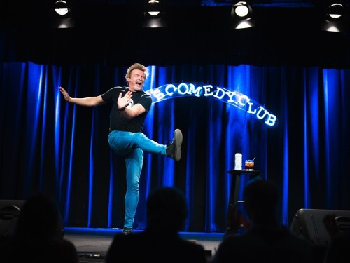 Another photo from Rhys's stand-up show. It looks like he is dancing, or maybe just showing off his legs, which are clad in a pair of very tight blue jeans. He is standing on one leg while kicking the other leg up towards the right, his arms stretched out towards the left for balance. His head is turned towards the right and his mouth is wide open in a laugh or shout.