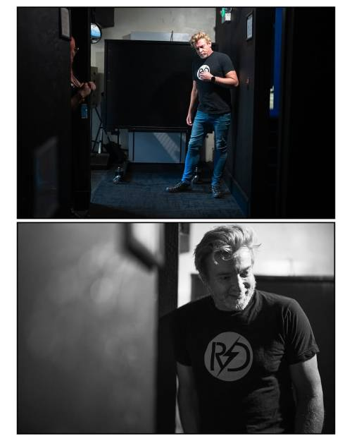 Two backstage photos of Rhys. In the one at the top, he is standing in front of a wall in a corridor or small room, leaning forward a little with one hand up near his chest and frowning slightly, probably in concentration as he prepares for his show. The photo at the bottom is in black and white, and we see Rhys from the hip up, leaning against a wall in a more relaxed posture. He is looking down and to the side with a smile on his face.