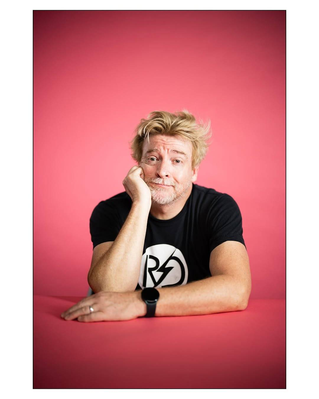 Portrait photo of Rhys, shot straight-on. He sits at a salmon pink table in front of a background of almost identical colour. He is propping up his head with his right hand, his elbow on the table, while his left forearm rests across the table top. He is looking directly at the camera with a smile, his eyebrows drawn up slightly in a soft, open expression. His blond hair looks artfully tousled, his ginger & white beard a little scruffy. He is wearing a black t-shirt with his RD logo printed on it in black & white.