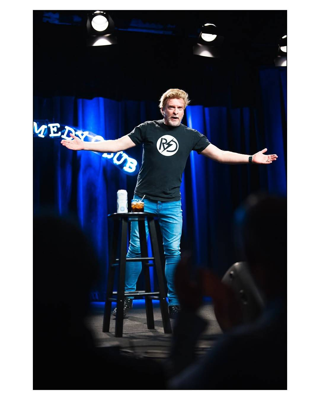 Photo from Rhys's stand-up show. He is standing behind the stool with the drinks now, leaning forward a little with his arms spread wide as he speaks.