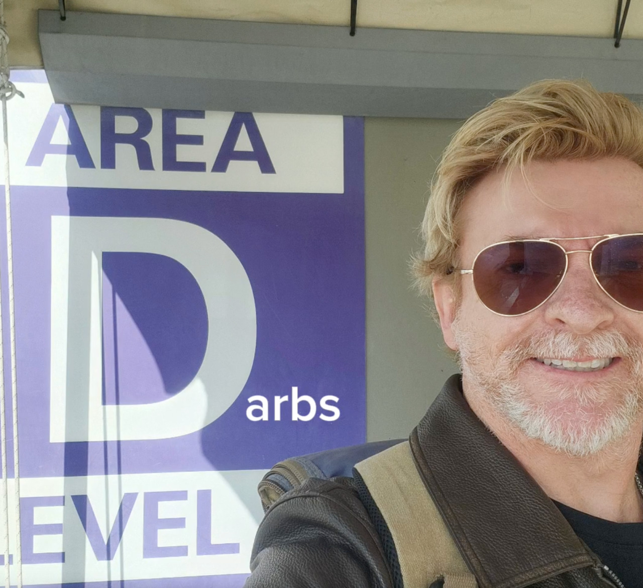 Selfie of Rhys Darby. He is standing in front of a sign that reads ‘Area D’ and is smiling at the camera. The photo has been edited to add ‘arbs’ after the D so that the sign reads ‘Area Darbs’.