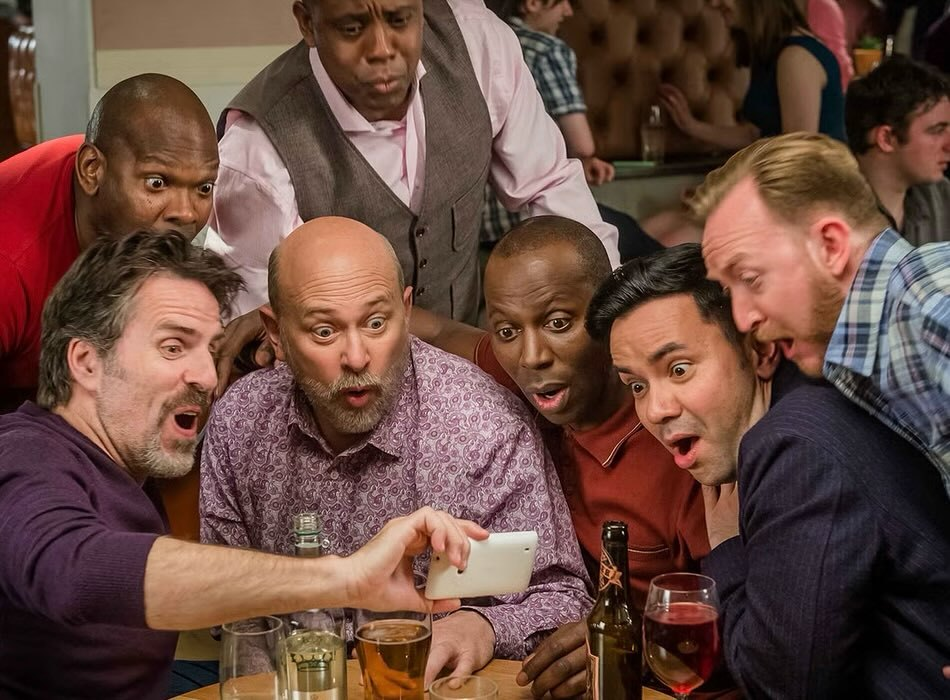 Production still from Cucumber: a group of 7 people, including Con’s character, look at a phone screen in surprise or shock, their eyes wide and mouths open.