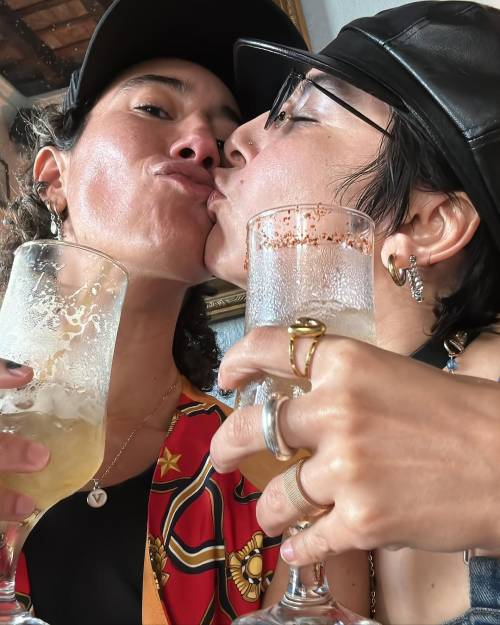 Selfie of Vico and one of their partners kissing. They are both seen from about the chest up, each holding a glass containing a pale, foamy drink, probably beer. Vico is looking down at the camera while their partner is seen in profile, kissing one corner of Vico’s mouth.