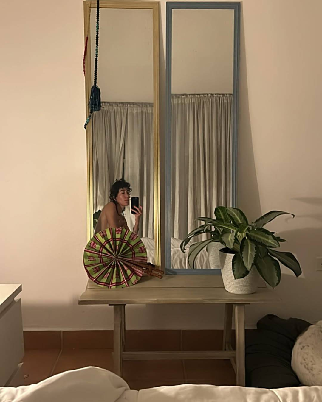 Another mirror selfie of Vico, this time sitting up in bed. Against the wall next to the bed stands a wooden table with 2 tall mirrors on top, and we see Vico’s reflection in one of them. Vico looks down at their phone screen as they take the photo. They appear to be topless, but their chest is covered by a round hand fan leaning against the mirror.