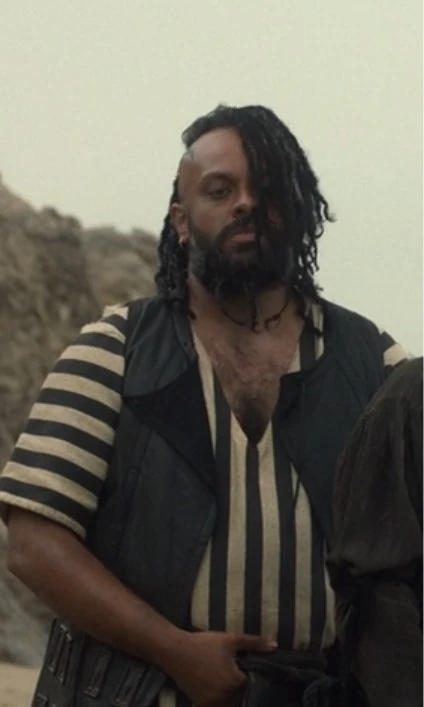 Guz Khan as Ivan in Our Flag Means Death season 1, standing with one hand on his belt. He is looking straight ahead and slightly down with a serious expression. His black dreadlocks fall down over one half of his face, and the other half of his head is shaved. He is wearing a black leather vest over a black & white striped t-shirt with a deep v-neck.