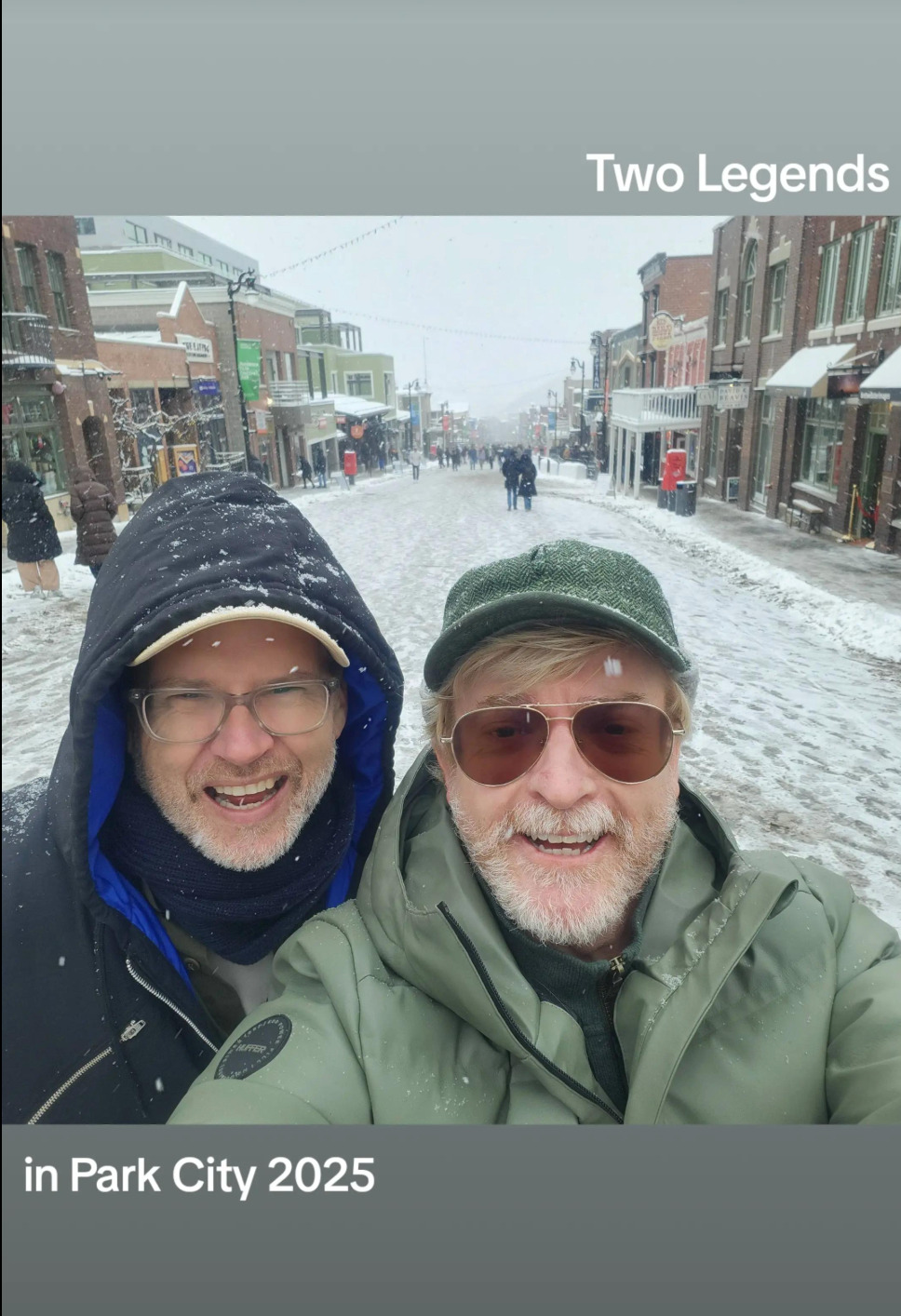 Selfie of Rhys and Buttons standing in the middle of a snow-covered street, both wearing winter coats, with Rhys also wearing a pair of aviator sunglasses and a tweed baseball cap. They smile broadly at the camera while snowflakes still fall around them. Text above the photo reads: 'Two Legends', and below: 'in Park City 2025'.