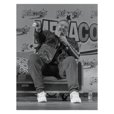 Black & white photo of Kristian on stage at MegaCon. He is holding up the microphone with one hand and pointing at someone in the audience with the other, smiling as he speaks.