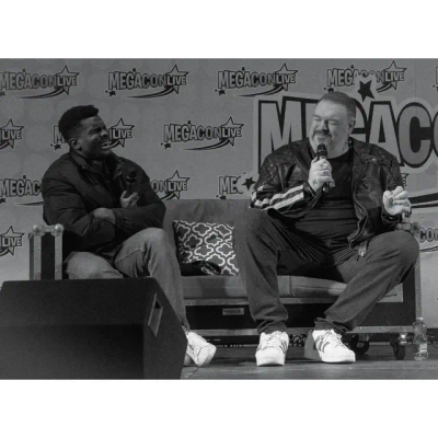 Black & white photo of Kristian and Clinton sitting on stage. Clinton is laughing at something Kristian said, while Kristian smiles back at him, still holding up the microphone.