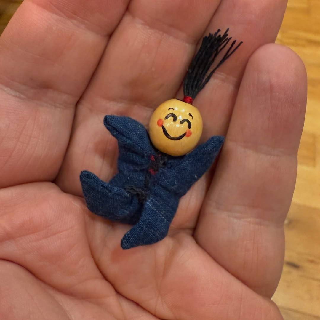 A small charm held in the palm of a hand. Its head is a yellow bead with a smiling face drawn on and a ponytail made from little pieces of black threads sticking out the top, and its body is made from tiny pieces of dark blue fabric sewn together in a kind of star shape to resemble arms and legs.