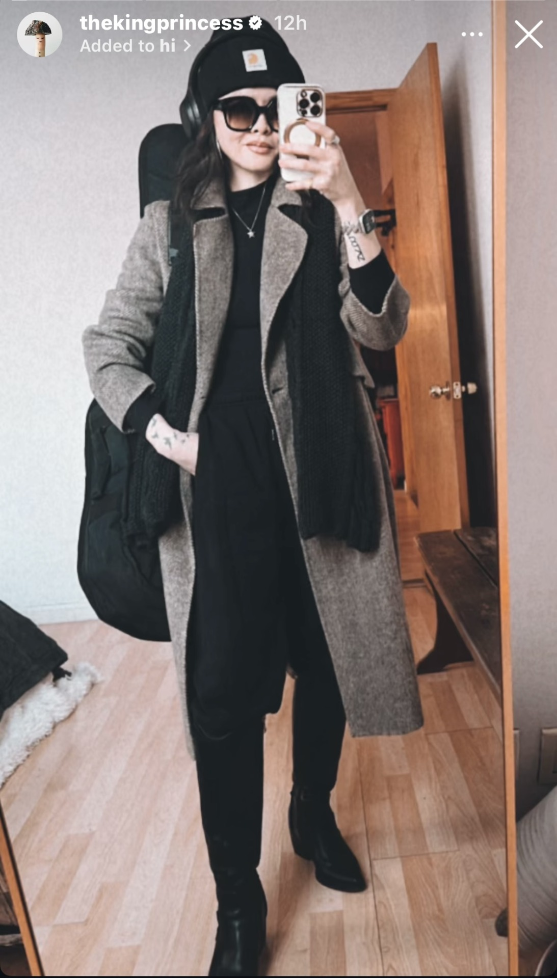 Mirror selfie from Ruibo's Instagram Stories. She is wearing black headphones over a black beanie, a large pair of sunglasses, and a grey winter coat over a black top paired with loose black trousers and black knee-high boots. Her black scarf is draped loosely over the top of her coat, and she is carrying a guitar case on her back.