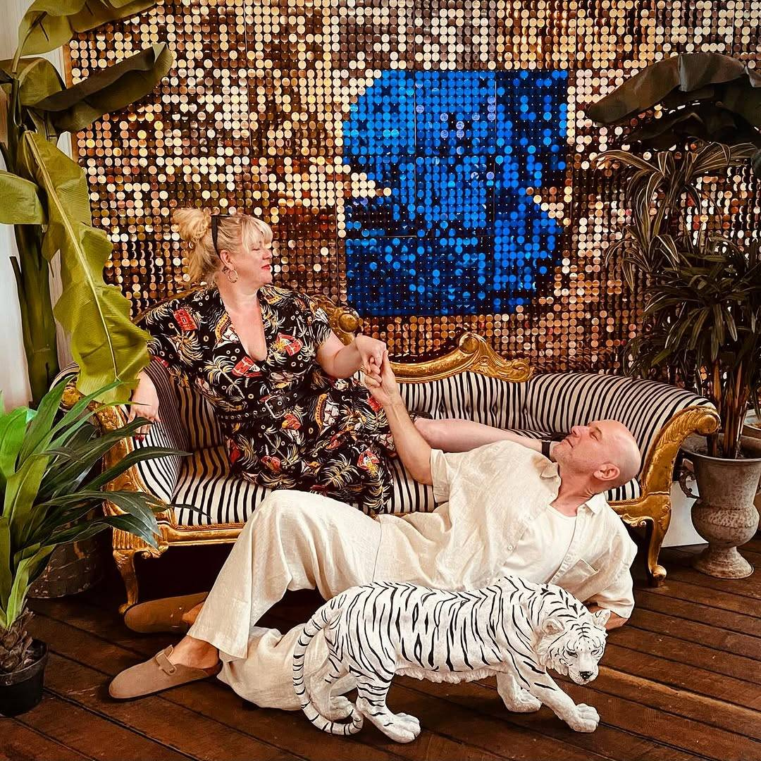 In front of a golden sequin wall featuring a large letter S made of blue sequins, Gypsy Taylor reclines on a zebra-striped chaise lounge with a golden frame, which is flanked by potted tropical plants of various sizes. Gypsy is wearing a black jumpsuit with a pattern of palm trees, sailing ships and treasure chests. She has her right elbow placed on the chaise lounge's arm rest, her left leg stretched out long and her right leg tucked under her. With a slight smile and her head held high, she looks down at a man dressed all in loose cream-coloured linen, who is lying on the wooden floor, propped up on one elbow and smiling back at Gypsy while holding her hand. In front of him, there is a small statue of a prowling white tiger.