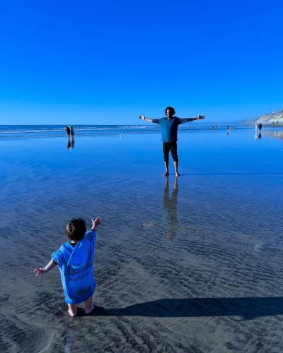 Beach photo of Samba and his kid, who is seen from behind and wearing a blue hoodie that reaches all the way down to their calves. The pair are standing in what looks like just a centimetre or so of water left behind by the tide, its smooth surface reflecting the clear blue sky. The child is reaching out to Samba, who stands a short distance away with his arms outstretched.